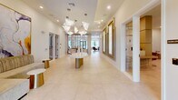 Located at 7430 Lyons Rd in Lake Worth, Florida, this thoughtfully designed single-floor commercial space offers a refined and welcoming environment across three distinct areas. The grand entrance lobby makes an immediate impression with expansive large-format cream tile flooring, soaring ceilings accented by a coffered tray detail, and dramatic sculptural pendant light fixtures in a frosted, organic form. Two striking backlit marble wall panels set within warm wood frames serve as a focal art feature, while a curved marble-and-gold center table anchors the open reception area with understated luxury. Recessed lighting throughout enhances the bright, airy atmosphere. The second area transitions into a warmly appointed lounge and waiting space, featuring light hardwood-style flooring, built-in wood shelving with accent lighting, and a teal-accented feature wall with horizontal banding. Comfortable upholstered seating, intimate booth-style banquettes, and tasteful contemporary artwork create a relaxed yet polished environment. Natural light filters in through a large window, lending a connection to the outdoors. A private consultation room offers a functional yet serene setting, complete with a wood-tone desk, guest seating, floor-to-ceiling glass overlooking a lush tropical landscape with palm trees, and an adjacent clinical or treatment alcove. The overall design balances elegance with functionality in a cohesive, upscale aesthetic.