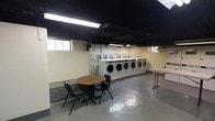 This single-floor laundry facility is designed with practicality and efficiency in mind. The room features a prominent row of commercial-grade front-load washing machines lined along one wall, each equipped with individual coin-operated or card payment units, making them well-suited for shared or high-volume use. A folding table positioned nearby provides a convenient surface for sorting and organizing laundry. The waiting area includes a round wooden table accompanied by two chairs, offering a comfortable spot for patrons while they wait. The space is illuminated by fluorescent ceiling lighting, which provides bright, even coverage throughout the room. The ceiling is painted black, giving the space an industrial character, while the walls are finished in a neutral cream tone. A painted concrete floor adds to the utilitarian aesthetic and ensures easy maintenance. Small windows along the upper portion of the walls allow for a modest amount of natural light and ventilation, complemented by a wall-mounted fan to support air circulation. The overall layout is open and efficiently organized, prioritizing ease of movement and access to all machines and surfaces.