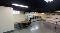 This single-floor laundry facility is designed with practicality and efficiency in mind. The room features a prominent row of commercial-grade front-load washing machines lined along one wall, each equipped with individual coin-operated or card payment units, making them well-suited for shared or high-volume use. A folding table positioned nearby provides a convenient surface for sorting and organizing laundry. The waiting area includes a round wooden table accompanied by two chairs, offering a comfortable spot for patrons while they wait. The space is illuminated by fluorescent ceiling lighting, which provides bright, even coverage throughout the room. The ceiling is painted black, giving the space an industrial character, while the walls are finished in a neutral cream tone. A painted concrete floor adds to the utilitarian aesthetic and ensures easy maintenance. Small windows along the upper portion of the walls allow for a modest amount of natural light and ventilation, complemented by a wall-mounted fan to support air circulation. The overall layout is open and efficiently organized, prioritizing ease of movement and access to all machines and surfaces.
