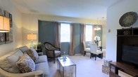This single-floor residence offers a thoughtfully designed open-concept layout that seamlessly connects the living and dining areas. The living room is anchored by plush, light-toned seating and complemented by an elegant glass coffee table, creating a refined yet comfortable atmosphere. A bold slate-blue accent wall serves as a striking focal point, adding depth and character to the space. Warm-toned drapes frame multiple windows that allow abundant natural light to fill the room, enhancing the airy and welcoming ambiance. Soft wall-to-wall carpeting runs throughout, adding warmth underfoot. The adjoining dining area is defined by a sleek dark table paired with crisp white upholstered chairs, centered beneath a classic chandelier that adds a touch of elegance. A floral arrangement on the dining table brings a fresh, inviting detail to the space. The overall color palette — combining cool blues, neutral creams, and warm accents — creates a balanced and harmonious environment. The layout flows intuitively, making the space feel both functional and stylish, well-suited for everyday living and casual entertaining alike.