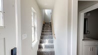 This three-story home offers a well-organized layout spanning three distinct floors, with three bedrooms, three full bathrooms, and a range of functional spaces designed for comfortable living. The ground floor houses an attached garage with an automatic opener, providing secure and convenient vehicle storage, alongside a dedicated laundry area. The main living level features an open-concept kitchen and living room, where crisp white shaker-style cabinetry, light countertops, and light wood-tone flooring establish a clean, modern aesthetic. Natural light filters throughout the home via large, multi-pane windows that frame outdoor views of surrounding greenery. The upper floor hosts the bedrooms, each finished with light hardwood flooring and neutral-toned walls. The primary bedroom offers a generous footprint with ample natural light. Ceiling fans are present in the bedrooms for added comfort. The bathrooms are finished with white subway tile walk-in showers featuring mosaic tile flooring, complemented by white vanities and clean hardware. Carpeted staircases with wooden handrails connect all three levels, while bright hallways with recessed lighting ensure smooth transitions throughout. The overall ambiance is contemporary, light-filled, and move-in ready.