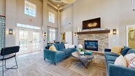 This single-story community clubhouse is designed to deliver both comfort and functionality across several distinct areas. The main lounge commands attention with its dramatic double-height ceilings and an abundance of natural light streaming through tall clerestory windows and multiple sets of French doors. Patterned carpet in neutral tones runs throughout, complementing the warm greige walls and bold teal upholstered seating. A striking floor-to-ceiling stacked stone fireplace with a wood mantel and wall-mounted television serves as the focal centerpiece of the lounge, while an elegant circular chandelier adds a refined finishing touch overhead. Adjacent to the lounge, a casual dining nook with white chairs and a dark wood table offers a quieter setting. A stylish barn door leads to a dedicated business center equipped with computer workstations and bar-height seating, ideal for remote work or study. A beverage and amenity station is conveniently positioned nearby. The fitness center, separated by glass-paned French doors, is generously outfitted with treadmills, ellipticals, a multi-station cable machine, a full dumbbell rack, weight benches, and floor-to-ceiling mirrors. Natural light fills the gym through multiple windows, and a ceiling fan ensures comfortable airflow during workouts. Together, these spaces create a polished, welcoming environment suited for both relaxation and productivity.
