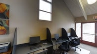 This single-floor computer lab offers a functional and focused environment designed for individual productivity. The space features a row of dedicated workstations, each equipped with a desktop computer, monitor, keyboard, and mouse, separated by dark privacy dividers that create a sense of personal space for each user. The workstations are arranged along a continuous desk surface, maximizing efficient use of the available area. A notable architectural feature is the angled, sloped ceiling that follows the roofline, lending the space a distinctive character. Tall, multi-pane windows allow natural light to filter in, brightening the warm, neutral-toned walls and contributing to a comfortable working atmosphere. The flooring features a patterned carpet, adding warmth underfoot. Ergonomic task chairs accompany each station, supporting extended use. The overall ambiance is utilitarian yet well-organized, making this space well-suited for a business center, library workroom, or shared office facility where users require reliable access to computing resources in a structured, distraction-minimized setting.