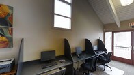 This single-floor computer lab offers a functional and focused environment designed for individual productivity. The space features a row of dedicated workstations, each equipped with a desktop computer, monitor, keyboard, and mouse, separated by dark privacy dividers that create a sense of personal space for each user. The workstations are arranged along a continuous desk surface, maximizing efficient use of the available area. A notable architectural feature is the angled, sloped ceiling that follows the roofline, lending the space a distinctive character. Tall, multi-pane windows allow natural light to filter in, brightening the warm, neutral-toned walls and contributing to a comfortable working atmosphere. The flooring features a patterned carpet, adding warmth underfoot. Ergonomic task chairs accompany each station, supporting extended use. The overall ambiance is utilitarian yet well-organized, making this space well-suited for a business center, library workroom, or shared office facility where users require reliable access to computing resources in a structured, distraction-minimized setting.