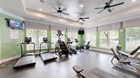 This fitness center in Annapolis, MD offers a comprehensive workout environment within a single, efficiently designed exercise room. The space is bathed in natural light streaming through large windows fitted with white roller shades and shutters, offering views of surrounding greenery. Sage green accent walls complement the crisp white tray ceilings, which are outfitted with multiple ceiling fans and recessed lighting to maintain a comfortable atmosphere during workouts. The durable speckled rubber flooring provides safe, cushioned support throughout the space. Cardio equipment includes treadmills, a StairMaster, ellipticals, and stationary bikes, all positioned along the windowed walls to take advantage of the outdoor views. A multi-station strength machine and a rack of dumbbells cater to resistance training needs, while stability balls add versatility for functional fitness. A full-length wall mirror expands the sense of space and aids in form correction. A bold, colorful word-art mural featuring fitness-themed typography serves as a dynamic focal point and motivational backdrop. A wall-mounted TV adds entertainment options during cardio sessions, rounding out this fully appointed community fitness facility.