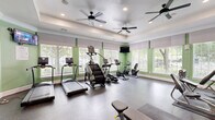 This fitness center in Annapolis, MD offers a comprehensive workout environment within a single, efficiently designed exercise room. The space is bathed in natural light streaming through large windows fitted with white roller shades and shutters, offering views of surrounding greenery. Sage green accent walls complement the crisp white tray ceilings, which are outfitted with multiple ceiling fans and recessed lighting to maintain a comfortable atmosphere during workouts. The durable speckled rubber flooring provides safe, cushioned support throughout the space. Cardio equipment includes treadmills, a StairMaster, ellipticals, and stationary bikes, all positioned along the windowed walls to take advantage of the outdoor views. A multi-station strength machine and a rack of dumbbells cater to resistance training needs, while stability balls add versatility for functional fitness. A full-length wall mirror expands the sense of space and aids in form correction. A bold, colorful word-art mural featuring fitness-themed typography serves as a dynamic focal point and motivational backdrop. A wall-mounted TV adds entertainment options during cardio sessions, rounding out this fully appointed community fitness facility.
