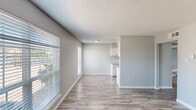 This single-floor unit presents a cohesive, updated interior finished throughout in light wood-look luxury vinyl plank flooring and soft gray paint, creating a bright and consistent aesthetic across all rooms. The open living and dining area is generously proportioned, with multiple large windows fitted with horizontal blinds that allow natural light to fill the space. A textured popcorn ceiling runs throughout, and clean white baseboards provide a crisp contrast to the neutral walls.

The living area connects directly to the kitchen, which is efficiently arranged in a galley-style layout. The kitchen features white upper and lower cabinetry with brushed nickel hardware, granite-look countertops, a stainless steel sink, a dishwasher, and a full-size refrigerator. A door within the kitchen provides access to additional storage or utility space.

The bedroom areas continue the same flooring and neutral color palette, offering well-sized rooms with ample wall space. One bedroom includes a ceiling-mounted HVAC vent and an adjacent bathroom accessible through a hallway. The entry area is defined by a solid white door and a large window providing views to the exterior. Overall, the unit presents a clean, move-in ready environment with consistent finishes throughout.