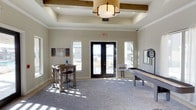 This single-floor amenity lounge is designed with a blend of modern and transitional styling, creating a welcoming, social atmosphere. The room's most notable architectural feature is its coffered ceiling, which incorporates rustic wood beam accents and a central pendant light fixture, adding warmth and visual interest overhead. Recessed lighting complements the natural light that streams in through flanking windows and a set of black-framed French doors, which open directly to an outdoor patio area, seamlessly connecting interior and exterior spaces.

The neutral, light gray walls and soft carpeted flooring establish a calm, versatile backdrop throughout the room. A high-top bistro table with industrial-style bar stools is positioned near the windows on one side, offering a casual spot for conversation or casual dining. On the opposite side, a full-length shuffleboard table serves as a central recreational feature, making the space well-suited for social gatherings and leisure activities. Wall art and a decorative mirror contribute subtle finishing touches. Overall, the lounge balances comfort, functionality, and style in a bright, airy setting.