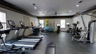 This fitness center is housed in a single-floor space designed to support a full range of exercise routines. The room features light-colored walls accented by a dark decorative border near the ceiling, lending the space a structured, purposeful aesthetic. Recessed lighting throughout the ceiling, complemented by a ceiling fan, ensures the room remains well-lit and comfortable during workouts.

The cardio section is prominently featured toward the front of the space, with multiple True-brand treadmills positioned for easy access. An elliptical trainer and stationary bikes, including a dedicated ride-style cycling unit with a display screen, round out the cardio offerings. A weight bench and additional resistance equipment are positioned along the perimeter to accommodate strength training.

A dedicated accessory wall organizes medicine balls, resistance bands, and exercise mats, keeping the space tidy and functional. Natural light filters in through several windows along the back wall, contributing to an open and energizing atmosphere. The dark rubber flooring provides durability and safety underfoot, making this fitness center a practical and well-appointed space for varied exercise needs.
