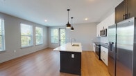 This single-floor residence showcases a contemporary open-concept design that seamlessly connects the kitchen and living area. The kitchen is a standout feature, equipped with stainless steel appliances including a refrigerator with ice maker, electric range, and over-the-range microwave. White shaker-style upper cabinets contrast with darker lower cabinets, complemented by a classic white subway tile backsplash and dark countertops along the perimeter. A central island with a white quartz countertop, undermount sink, and matte black pendant lighting overhead serves as a functional and stylistic focal point.

The open living area benefits from abundant natural light streaming through multiple large windows with views of surrounding greenery, and a sliding door provides additional access and light. Warm hardwood-style flooring runs consistently throughout the living spaces.

The bathroom features dark large-format floor tiles, a white vanity with grey countertop, and a tub/shower combination lined with gleaming white subway tiles and matte black fixtures, lending a clean, modern aesthetic.

A generously sized walk-in closet is outfitted with multi-level wire shelving and hanging rods on three walls, offering versatile storage throughout.