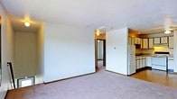 This single-floor space features an open living area finished with wall-to-wall carpeting in a neutral tone, providing a comfortable and cohesive foundation throughout the main room. The walls are painted white, contributing to a clean, bright atmosphere, while dark wood baseboards add a subtle contrasting detail along the perimeter. The textured ceiling adds modest visual interest overhead.

Adjacent to the living area, a partial view into the kitchen reveals white cabinetry and a white refrigerator, suggesting a practical galley-style kitchen layout. The kitchen area transitions naturally from the main living space, making it easily accessible.

A hallway visible from the living room leads further into the unit, with a ceiling-mounted light fixture providing illumination along the corridor. The layout appears straightforward and efficient, with clear sightlines connecting the living area, kitchen, and hallway. Overall, the space presents a functional, no-frills interior suited to everyday living, with neutral finishes that provide a versatile backdrop for personalization.
