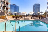 Located at 2333 Calle Del Mundo in Santa Clara, CA, this thoughtfully designed outdoor amenity space serves as a vibrant communal hub set within a modern multi-story residential complex. The courtyard is anchored by a large, resort-style swimming pool with crystal-clear water, flanked by sun loungers and market umbrellas that invite relaxation under the California sun. Surrounding the pool, wide-format porcelain tile pavers create a clean, contemporary aesthetic throughout the space.

Raised planter beds constructed of warm sandstone-toned masonry introduce lush, drought-tolerant landscaping, adding greenery and natural texture to the hardscape. A built-in outdoor kitchen station, equipped with a stainless steel gas grill and ample counter space, provides an ideal setup for al fresco entertaining. Multiple seating zones are thoughtfully arranged across the courtyard, including a generous sectional lounge area with plush neutral cushions and woven accent chairs perfect for gathering.

A rooftop terrace area offers elevated views of the Santa Clara skyline, including neighboring high-rise towers, with comfortable lounge seating arranged around a linear fire feature. Glass-paneled railings maximize the open sightlines. The space is framed by the building's modern façade, characterized by a mix of concrete, warm corten-style metal cladding, and large-paned balcony windows throughout.