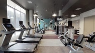 This fitness center offers a comprehensive workout environment designed with both functionality and aesthetics in mind. The space is organized into distinct workout zones, each catering to different training needs. A generously sized cardio area lines the windowed perimeter, housing a full row of treadmills, elliptical machines, and stationary bikes, all positioned to take advantage of the abundant natural light streaming through large floor-to-ceiling windows with views of the surrounding greenery. Mounted flat-screen televisions provide entertainment during workouts. The strength training zone features a well-stocked free weight section with a full dumbbell rack, weight benches, and a multi-functional cable and resistance machine system. Floor-to-ceiling mirrors amplify the sense of space and allow users to monitor their form. Stability balls and additional accessories round out the functional fitness offerings. The design palette blends cool teal accent walls with a dark, recessed ceiling punctuated by stylish pendant lighting and exposed concrete columns, lending an industrial-chic ambiance. A dedicated amenity counter near the entrance, finished with a granite surface, a beverage cooler, and a tiled backsplash, provides towel service and refreshments. Private rooms off the main corridor offer additional facilities, completing this full-service fitness experience.