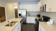 This single-floor residence offers a well-appointed, open-concept layout designed for comfortable everyday living. The kitchen serves as a central highlight, showcasing crisp white cabinetry extending to the ceiling for ample storage, speckled granite countertops, and a full suite of stainless steel appliances including a French door refrigerator with ice maker, electric range, built-in microwave, and dishwasher. A generous island with an undermount double sink, pendant lighting overhead, and barstool seating makes the kitchen equally suited for meal preparation and casual dining. Dark hardwood-style flooring runs throughout, providing a warm contrast to the light cabinetry and countertops.

The open living and dining area flows naturally from the kitchen, offering room for a full dining table and a comfortable seating arrangement. Large sliding glass doors flood the space with natural light and provide direct access to an outdoor patio, where views of surrounding greenery enhance the overall ambiance. The bedroom, visible from the living area, is conveniently positioned for easy access, completing this cohesive and functional single-level layout.