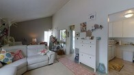 This single-floor space features a welcoming living area characterized by its striking vaulted ceilings, which create an impressive sense of height and openness. The walls are finished in a clean white and soft gray palette, amplifying the natural light that streams in through multiple windows and a glass door, lending the space an airy, bright atmosphere.

The living area is anchored by a white leather sofa adorned with a decorative starfish pillow, reinforcing the coastal theme carried throughout the space. A patterned coral and white area rug adds warmth underfoot. A large mirror near the entrance enhances the sense of depth, reflecting light and greenery from outside.

A white storage cabinet with multiple drawers serves as both a functional and decorative element along one wall, topped with coastal-themed ornaments including coral pieces and decorative figures. Framed artwork and beach-inspired prints contribute to the nautical ambiance.

Natural light is a defining feature, with windows and a glazed door offering views of surrounding greenery. The overall aesthetic is cohesive, relaxed, and effortlessly tied to a breezy coastal theme.