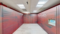 The Mail Room at Arrive Stamford is a dedicated, single-floor package management facility designed to streamline the delivery and retrieval experience for residents. The room is defined by floor-to-ceiling automated parcel lockers that line three walls, wrapped in a bold, branded graphic treatment in deep reds and geometric patterns, prominently displaying the Arrive Stamford logo. The lockers vary in size to accommodate packages of different dimensions, ensuring versatility for all types of deliveries. An integrated electronic kiosk with a touchscreen interface and keyboard provides residents with a convenient, self-service retrieval system. The space features light wood-look flooring that contrasts cleanly with the vivid locker panels, while a drop ceiling with recessed panel lighting ensures the room is well-lit and functional. A secure staff-access door is also present, supporting operational management of the space. The overall design is clean, modern, and on-brand, balancing practicality with a polished aesthetic that reflects the Arrive Stamford community's attention to resident convenience.