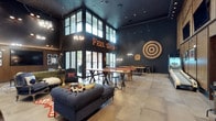 This single-level clubroom makes a strong first impression with its dramatic double-height ceilings and deep, dark color palette of navy and charcoal tones that lend a sophisticated, moody ambiance. Large floor-to-ceiling windows on one wall flood the space with natural light, offering views to the outdoors while contrasting beautifully against the dark interior. A illuminated marquee sign reading "Feel Good" serves as a standout focal point, reinforcing the room's energetic, social character. The expansive layout is thoughtfully divided into distinct zones for relaxation and recreation. A plush, tufted navy sofa anchors the lounge area, complemented by accent chairs and layered rugs that add warmth and texture. The game area features a pool table alongside high communal tables with industrial-style seating, ideal for gathering and socializing. A large bullseye target mounted on the wall adds a playful, decorative touch. Pendant lighting and wall-mounted fixtures provide warm, layered illumination throughout. Large-format floor tiles ground the space and enhance its open, airy feel. Overall, the clubroom balances bold design with functional social spaces, making it an inviting hub for leisure and community.