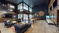This single-level clubroom makes a strong first impression with its dramatic double-height ceilings and deep, dark color palette of navy and charcoal tones that lend a sophisticated, moody ambiance. Large floor-to-ceiling windows on one wall flood the space with natural light, offering views to the outdoors while contrasting beautifully against the dark interior. A illuminated marquee sign reading "Feel Good" serves as a standout focal point, reinforcing the room's energetic, social character. The expansive layout is thoughtfully divided into distinct zones for relaxation and recreation. A plush, tufted navy sofa anchors the lounge area, complemented by accent chairs and layered rugs that add warmth and texture. The game area features a pool table alongside high communal tables with industrial-style seating, ideal for gathering and socializing. A large bullseye target mounted on the wall adds a playful, decorative touch. Pendant lighting and wall-mounted fixtures provide warm, layered illumination throughout. Large-format floor tiles ground the space and enhance its open, airy feel. Overall, the clubroom balances bold design with functional social spaces, making it an inviting hub for leisure and community.
