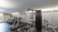 This well-equipped fitness center at 10463 Corbeil Dr in Saint Louis, Missouri offers a comprehensive workout environment within a single-floor layout. The space features dark rubber flooring throughout, providing a durable and functional surface suited for a variety of exercise activities. Large windows along one wall allow natural light to fill the room and offer views of an outdoor courtyard area, creating a bright and welcoming atmosphere.

The cardio section is outfitted with multiple Matrix treadmills, elliptical trainers, and a stationary bike, arranged in a row to maximize space and allow for easy use. Adjacent to the cardio equipment is a multi-station cable strength machine, accompanied by a free-weight dumbbell rack and a workout bench, catering to a range of strength training needs. Instructional exercise posters are displayed on the walls for user reference.

A wall-mounted flat-screen television is positioned for viewing during workouts. The facility also includes a sauna room, adding a wellness and recovery component to the space. The entry area features tile flooring, a glass-paned door, and a wall-mounted air conditioning unit, ensuring a comfortable environment year-round. Recessed and overhead lighting supplements the natural light, keeping the space well-lit at all hours.