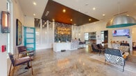 Located at 219 Depot Ave in Delray Beach, Florida, Delray Station presents a thoughtfully designed single-floor space that combines contemporary aesthetics with a welcoming, community-oriented atmosphere. The lobby serves as the centerpiece of the space, immediately drawing attention with its dramatic mosaic tile accent wall emblazoned with the illuminated "Delray Station" marquee sign — a defining architectural feature that sets the tone for the entire environment.

The reception area features a sleek white front desk complemented by a mosaic-clad service counter, providing a polished and professional point of contact. Polished concrete floors extend throughout, lending an industrial-chic character that ties the space together cohesively. Soaring ceilings amplify the sense of openness, while a suspended bronze dome pendant light anchors the lounge area with a bold decorative statement.

The lounge is furnished with an eclectic mix of mid-century modern seating — including tufted leather chairs, patterned accent chairs, and a low coffee table — arranged to encourage conversation and comfort. Large windows and glass doors flood the interior with abundant natural light and offer views of the surrounding greenery. A wall-mounted flat-screen TV and a display console add functional and entertainment elements to the space. Teal accent doors throughout provide vibrant pops of color, reinforcing a distinctive and cohesive design identity.