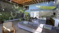 This single-floor space showcases a striking blend of industrial and contemporary design elements. The soaring exposed ceiling reveals raw wooden beams, metal ductwork, and pendant lighting suspended from above, creating an open, loft-like atmosphere with impressive vertical volume. Polished concrete floors run throughout, lending a sleek, modern foundation to the interior.

A bold, large-scale green patterned accent wall serves as a dramatic focal point, adding artistic depth and vibrancy to the space. Complementing this, a textured stone or tile feature wall on the opposite side introduces warmth and contrast. The interior is organized into distinct functional zones: a lounge area furnished with a generous striped sectional sofa and accent pillows anchors one section, while a dining or meeting area with green chairs and glass-top tables occupies another. A counter-height bar or reception area separates zones with practical elegance.

Abundant natural light pours in through large windows and glass doors, illuminating the space and connecting it visually to a landscaped outdoor area. Greenery and decorative branches add organic touches throughout, enhancing the overall fresh, modern ambiance.