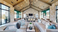 Located at 18131 Bluegrass Park Ln in Cypress, TX, this beautifully designed single-story clubhouse is a standout gathering space that masterfully blends rustic charm with modern elegance. The architecture is defined by dramatic vaulted ceilings with exposed wooden beams and light-toned wood planking overhead, creating an expansive, airy atmosphere throughout the open-concept interior.

The main living area is anchored by a generous seating arrangement featuring a plush sofa, swivel accent chairs in soft teal, and a glass-topped coffee table, all set atop a patterned area rug. Striking sculptural chandeliers and pendant lighting add a refined, contemporary touch to the soaring ceiling above.

Adjacent to the lounge is a formal dining area with a sleek, dark dining table surrounded by mixed seating, bathed in natural light streaming through large floor-to-ceiling windows that offer views of the surrounding community. Teal accent walls and teal-framed windows punctuate the neutral stone and wood palette, adding vibrant pops of color throughout.

The kitchen area features a large island with white countertops, weathered wood cabinetry, woven bar stools, and patterned tile flooring. A full wall of stacked stone provides a dramatic backdrop behind the counter and wall-mounted TV. Private meeting rooms with glass-paneled doors offer additional flexible space, completing this well-appointed community destination.