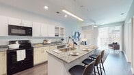 This well-appointed single-floor residence offers a thoughtfully designed open-concept layout that seamlessly connects the kitchen, dining, and living areas. The kitchen serves as the centerpiece of the home, showcasing granite countertops, a large central island with bar seating, ample white cabinetry with open shelving, and black appliances including a refrigerator, range, microwave, and dishwasher. Pendant and track lighting illuminate the space, while wood-style flooring extends throughout the main living areas, adding warmth and continuity.

The living area is bright and inviting, benefiting from natural light through large windows and a ceiling fan that enhances airflow and comfort. The bedrooms are generously proportioned, featuring plush carpeting, ceiling fans, and large windows that invite natural light. Each bedroom includes a walk-in closet with built-in shelving and hanging rods, offering excellent storage capacity.

The bathroom is finished with large-format marble-look wall tile in the walk-in shower, complemented by wood-tone floor tile, a granite-topped vanity, and clean, neutral tones throughout. The overall aesthetic of the residence is modern and cohesive, characterized by a crisp white and grey palette, quality finishes, and a functional flow that makes everyday living both comfortable and stylish.