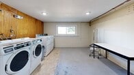 Located at 78 Laning St, this shared laundry facility offers a practical and well-organized space designed to accommodate the laundry needs of multiple users. The single-floor layout provides an open, spacious feel with a concrete floor throughout, offering durability and ease of maintenance.

The room is lined with warm wood-paneled walls on two sides, lending a degree of character to the otherwise utilitarian space. A row of commercial-grade front-loading washers and dryers lines one wall, providing ample capacity for heavy-use laundering. A large, multi-tiered folding and sorting table runs along the wood-paneled wall, offering generous surface and shelf space for organizing laundry. An additional folding table with a chair is positioned in the open area, providing a convenient spot for sorting or waiting.

Natural light enters through a sliding window on one wall, supplemented by recessed ceiling lighting that keeps the room well-lit. Exposed utility piping runs along the ceiling, reflecting the room's functional design. Posted laundry instructions are visible near the machines, ensuring users can operate the equipment correctly. A rolling laundry cart is also available for added convenience. The space is straightforward, clean, and purpose-built for efficient shared laundry use.