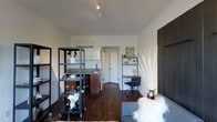 This single-floor space showcases a thoughtfully arranged open-plan layout that maximizes functionality within a compact footprint. The interior features rich dark hardwood flooring throughout, which contrasts sharply with the clean white walls and ceiling, lending the space a modern, high-contrast aesthetic.

A kitchenette occupies one side of the space, equipped with granite countertops, a undermount sink, warm wood cabinetry, and white upper cabinets. A wall-mounted mini-split HVAC unit ensures climate control. Adjacent to the kitchen, a freestanding black metal open shelving unit on casters serves as both a room divider and a display and storage solution, styled with glassware, botanicals, and decorative objects, adding an industrial touch.

A dedicated workspace is positioned along one wall, featuring a compact desk with a rolling office chair and a wall-mounted shelf displaying decorative accents. Nearby, a tall decorative branch arrangement in a metallic vase adds a sculptural element.

The living area anchors the far end of the room, with a sectional sofa partially visible. Track lighting on the ceiling provides directed illumination throughout. A doorway leads to an additional room, suggesting further private space beyond the open-plan area.