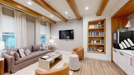 Located at 9000 Palm Passage Loop in Summerville, SC, this thoughtfully designed single-floor amenity space offers a versatile and inviting environment for both relaxation and productivity. The space is defined by its striking exposed wood ceiling beams, which run throughout and lend a warm, contemporary character to the neutral, light-toned interiors. Light wood plank flooring flows seamlessly from room to room, reinforcing a cohesive and welcoming aesthetic.

The main lounge area features a comfortable seating arrangement with a plush sofa, cognac leather accent chairs, and a central coffee table, anchored by a wall-mounted flat-screen TV. A built-in wood-framed bookshelf adds both function and visual interest. Natural light filters in through large windows dressed with sheer linen curtains, brightening the space considerably.

Adjacent to the lounge is a well-appointed café-style seating area with banquette seating set against a slatted wood accent wall, illuminated by oversized pendant lights. Round white café tables and wooden chairs create a casual gathering spot. A dedicated coffee and print station with white cabinetry provides everyday convenience.

Private individual workstations, fully enclosed in warm wood slatted panels, offer quiet, focused spaces equipped with desktop computers — ideal for residents seeking a more concentrated work environment.