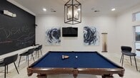 Located at Madison Square Place in Charlotte, NC, this amenity space at The District delivers a well-appointed community lounge designed for both leisure and social engagement. The main gathering area is anchored by a full-size billiard table dressed in deep blue felt, positioned beneath a striking geometric lantern-style chandelier that provides warm, focused lighting. White tile flooring runs throughout, complementing the clean, bright walls adorned with bold abstract paintings in navy and charcoal tones, adding an artistic energy to the room. A large-format chalkboard accent wall serves as both a design feature and a dynamic community board, flanked by bar-height seating that creates an informal gathering spot. A wall-mounted flat-screen television completes the entertainment setup. Adjacent to the main lounge, a cozy seating nook with large windows frames views of the lush outdoor surroundings, filling the space with natural light and offering a tranquil retreat for conversation or quiet moments. A grand staircase with dark wood newel posts, rich wood handrails, and sleek black iron balusters connects to upper levels, adding architectural distinction to the overall design. The space balances refined aesthetics with functional, community-centered amenities throughout.