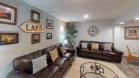 Located at 2400 Interlachen Rd in Spring Park, Minnesota, this single-floor space offers a well-organized and inviting layout designed for comfortable gathering and relaxation near the lake. The open main area features distinctive multi-toned slate tile flooring that flows throughout, anchoring the space with a grounded, rustic character. Structural columns define zones within the open floor plan without closing off the space, maintaining an airy and connected feel throughout.

The lounge area is appointed with rich dark leather sofas arranged around a woven area rug, complemented by lake-themed wall décor including framed photography and a bold "LAKE" directional sign. A large flat-screen television and a navy blue console add functionality and style to this relaxed seating area.

The dining section accommodates multiple round wood tables with slat-back chairs, providing generous seating for groups. A built-in bar counter with dark stone countertops runs along one wall, backed by pass-through windows into the kitchenette, offering a casual and social dining experience.

The kitchenette is efficiently designed with warm oak cabinetry, dark countertops, a double sink, microwave, and compact appliances set on light ceramic tile flooring. Recessed lighting throughout the space ensures consistent, practical illumination, while sheer curtains soften natural light from exterior-facing windows.