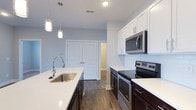 Located at 6770 Printers Blvd in New Albany, Ohio, this single-floor residence offers a thoughtfully designed living space with contemporary finishes throughout. The kitchen serves as a standout feature, showcasing a sleek, galley-style layout with crisp white shaker cabinetry that extends to the ceiling, providing generous storage. Rich, dark hardwood-style flooring creates a striking contrast against the bright white quartz countertops, lending the space a polished, modern aesthetic. Stainless steel appliances — including a range, microwave, and dishwasher — are seamlessly integrated into the design, elevating both form and function. A deep single-basin stainless steel sink with a high-arc gooseneck faucet anchors the kitchen island, which offers additional workspace and casual seating potential. Stylish pendant lighting hangs above the island, adding warmth and visual interest. Soft, neutral-toned walls and recessed lighting keep the interior feeling bright and airy. A set of double doors hints at generous closet or storage space nearby. The overall ambiance is clean, contemporary, and move-in ready, making this New Albany residence an appealing option for those seeking modern comfort and refined design.