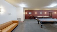 Located at 739 S 300 W in Salt Lake City, Utah, this recreational amenity space offers a thoughtfully designed game room complemented by a connecting corridor. The game room is defined by a bold deep-red accent wall that anchors the space, contrasting with crisp white walls on the opposing side. Soft wall-mounted sconces provide warm, ambient lighting throughout, supplemented by flush ceiling fixtures that illuminate the textured ceiling above. The room is carpeted in a neutral gray, lending a comfortable and cohesive feel underfoot. A full-size billiard table serves as the centerpiece, positioned prominently in the open floor plan, while a regulation shuffleboard table runs along the feature wall. A foosball table and a high-top bar table with stools offer additional social and recreational options. A leather sofa provides a comfortable lounge area for spectators. Framed artwork on the accent wall adds a finishing decorative touch. The adjoining corridor is clean and well-lit, featuring the same gray carpet, white walls, warm wood-trimmed doors, and wall sconces that create a welcoming transition through the building. Together, these spaces form a functional and inviting communal area well-suited for leisure and social gatherings.