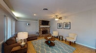 This single-floor clubhouse offers a well-appointed and versatile gathering space designed to accommodate a variety of community functions. The main lounge area serves as the centerpiece of the space, anchored by a striking full-brick fireplace with a white mantel and a wall-mounted flat-screen television above it. Comfortable seating, including a plush sofa, accent chairs, and a coffee table atop a teal geometric area rug, creates a relaxed and inviting atmosphere. Ceiling fans throughout the space ensure year-round comfort. Natural light flows generously through large windows fitted with white blinds and a set of French doors, brightening the tile-floored interior and creating an airy feel. Adjacent to the lounge, an open dining area accommodates a wood dining table with mixed seating, making it well-suited for meetings or shared meals. Decorative artwork adorns the soft gray walls, adding a polished touch. The kitchenette, located at one end of the space, features white cabinetry, a full-size refrigerator, a microwave, and counter space for food preparation and serving. A secondary entry door provides convenient access from an exterior area. The open, flowing layout of the clubhouse makes it an adaptable and functional venue for the community.