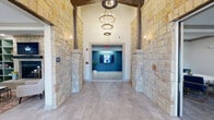 This single-floor professional space combines rustic architectural character with contemporary interior design to create a polished and welcoming environment. The grand entry corridor immediately sets the tone, featuring floor-to-ceiling natural limestone-clad walls, exposed dark wood beam accents, and a striking two-tiered wrought-iron chandelier suspended beneath soaring vaulted ceilings. Large-format neutral tile flooring flows throughout the entry, creating a seamless transition into the interior.

Beyond the entry, the reception area offers a sleek, dark wood desk accompanied by modern navy blue lounge chairs with gold-toned frames, set against plush textured carpet. Multiple windows allow natural light to filter in, framing views of the surrounding exterior stonework. A private conference room features a round marble-topped table surrounded by coordinating navy chairs beneath a clean, coffered ceiling.

A comfortable lounge area showcases a stone-surround fireplace flanked by built-in sage green shelving units filled with books and décor, anchored by a leather sofa and accent chairs. A kitchenette area includes sage green cabinetry, a beverage refrigerator, and a sink, accompanied by a casual seating arrangement and access to resident mailboxes. Throughout, neutral warm tones, curated artwork, and abundant greenery contribute to a professional yet inviting ambiance.
