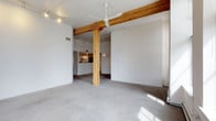 This single-floor space showcases an industrial-inspired aesthetic through a thoughtful combination of raw and refined materials. The open-concept layout is anchored by a striking exposed wood structural column and matching overhead beam, which add warmth and character against the predominantly white walls and ceiling. Concrete flooring runs throughout, contributing to the loft-like atmosphere while providing a durable, low-maintenance surface. A ceiling fan is centrally mounted, offering both practical airflow and a subtle design element. Track lighting is installed along the ceiling, allowing for flexible and adjustable illumination across the space. Toward the rear, a kitchen area is partially visible, featuring a raised counter or bar-height surface and standard appliances, separated from the main living area by a subtle change in flooring material. The white walls serve as a neutral canvas, making the space highly adaptable for various furnishing styles. Natural light filters in, casting soft shadows across the floor and enhancing the open, airy feel of the interior.