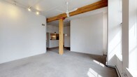 This single-floor space showcases an industrial-inspired aesthetic through a thoughtful combination of raw and refined materials. The open-concept layout is anchored by a striking exposed wood structural column and matching overhead beam, which add warmth and character against the predominantly white walls and ceiling. Concrete flooring runs throughout, contributing to the loft-like atmosphere while providing a durable, low-maintenance surface. A ceiling fan is centrally mounted, offering both practical airflow and a subtle design element. Track lighting is installed along the ceiling, allowing for flexible and adjustable illumination across the space. Toward the rear, a kitchen area is partially visible, featuring a raised counter or bar-height surface and standard appliances, separated from the main living area by a subtle change in flooring material. The white walls serve as a neutral canvas, making the space highly adaptable for various furnishing styles. Natural light filters in, casting soft shadows across the floor and enhancing the open, airy feel of the interior.