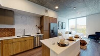 This contemporary single-floor residence showcases an industrial-modern aesthetic defined by striking exposed concrete ceilings throughout, warm wood-tone flooring, and an abundance of natural light. The open-concept kitchen is outfitted with stainless steel appliances, including a gas range, dishwasher, and French door refrigerator, complemented by warm wood cabinetry, a marble-style tile backsplash, and a white quartz island with seating — ideal for both cooking and casual dining. The living area flows seamlessly from the kitchen and is anchored by a large floor-to-ceiling window offering broad outdoor views, creating an airy, light-filled atmosphere. A dedicated sitting nook features an exposed concrete column, hardwood flooring, and ample wall space for art display. The bedroom is generously proportioned, featuring a ceiling fan, oversized windows with outdoor views, and room for a full furniture arrangement. A walk-in closet with built-in shelving and organization systems also houses a stacked in-unit washer and dryer for added convenience. The bathroom is finished with a floating wood-framed vanity, white quartz countertop, large mirror, and wood-look tile flooring, reflecting a clean, modern sensibility throughout.
