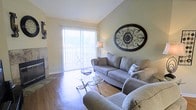 This single-story space offers a thoughtfully designed living area that combines comfort and style. The living room immediately draws attention with its vaulted ceilings, which lend an open, airy feel to the space. Warm hardwood flooring runs throughout, adding a sense of continuity and natural warmth to the room.

A stone-faced fireplace anchors one wall, providing both a functional heating element and a classic focal point. On the opposite side, a large sliding glass door fitted with vertical blinds allows generous natural light to fill the room, creating a bright and inviting atmosphere while offering access to the outdoors.

The living area is generously proportioned, comfortably accommodating multiple seating arrangements. A decorative oval wrought-iron wall piece adds an elegant, artistic touch to the neutral-toned walls, complementing the soft, neutral color palette throughout. A glass-top coffee table with a metal frame adds a contemporary accent to the space.

Overall, the living area strikes a balance between classic and transitional design, offering a comfortable and well-lit environment suited for both relaxing and entertaining.