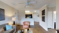 This single-floor residence offers an open-concept layout that seamlessly connects the living area to the kitchen. The living space features rich hardwood-style flooring that extends throughout, creating a warm and cohesive aesthetic. Crown molding frames the ceiling, adding a refined architectural detail, while a ceiling fan with a light fixture provides both airflow and ambient lighting.

The kitchen is positioned behind a raised peninsula counter, offering a natural division between the living and cooking areas without sacrificing the open feel. Dark cabinetry contrasts with the light-colored walls, and black appliances are visible within the kitchen space. The peninsula serves as a functional transition point, ideal for casual dining or food preparation.

The living area is generously proportioned, with neutral gray walls that provide a versatile backdrop. Decorative wall art adds visual interest to the space. Multiple doorways are visible, suggesting easy access to additional rooms. Overall, the space presents a balanced combination of functional design and comfortable living, with consistent finishes throughout contributing to a unified and inviting ambiance.