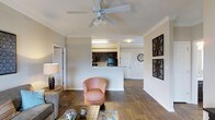 This single-floor residence offers an open-concept layout that seamlessly connects the living area to the kitchen. The living space features rich hardwood-style flooring that extends throughout, creating a warm and cohesive aesthetic. Crown molding frames the ceiling, adding a refined architectural detail, while a ceiling fan with a light fixture provides both airflow and ambient lighting.

The kitchen is positioned behind a raised peninsula counter, offering a natural division between the living and cooking areas without sacrificing the open feel. Dark cabinetry contrasts with the light-colored walls, and black appliances are visible within the kitchen space. The peninsula serves as a functional transition point, ideal for casual dining or food preparation.

The living area is generously proportioned, with neutral gray walls that provide a versatile backdrop. Decorative wall art adds visual interest to the space. Multiple doorways are visible, suggesting easy access to additional rooms. Overall, the space presents a balanced combination of functional design and comfortable living, with consistent finishes throughout contributing to a unified and inviting ambiance.