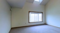 This two-floor property offers a thoughtfully designed layout that blends comfort and functionality. One of its most striking features is the soaring vaulted ceiling, which creates an impressive sense of openness and volume. Dual skylights are positioned overhead, flooding the interior with abundant natural light throughout the day and enhancing the airy feel of the space. A large bay window with warm wood trim serves as a prominent architectural detail, framing outdoor views and drawing even more light into the room. The neutral, cream-toned walls provide a clean and versatile backdrop, complementing the soft, wall-to-wall carpeting that adds warmth underfoot. The combination of the elevated ceiling, skylights, and generous window proportions gives the space a welcoming and expansive character. The two-floor layout offers distinct living zones, providing both shared and private areas suited to a variety of lifestyles. Overall, this property presents a bright, well-lit interior with classic architectural details that contribute to a timeless and inviting ambiance throughout.