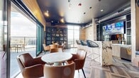 This sophisticated lounge space seamlessly blends indoor comfort with outdoor living across a well-appointed single floor. Inside, the interior design is anchored by striking herringbone-patterned hardwood flooring and warm wood-paneled walls that create a rich, contemporary aesthetic. A bold matte black ceiling with exposed industrial ductwork and track lighting adds an urban edge, complementing pendant fixtures that illuminate the space with a moody glow.

The centerpiece of the interior is a dramatic marble-faced bar island with veined white-and-gold stone, paired with sleek cabinetry, a wet bar with a sink, and a built-in beverage cooler. Bar-height woven chairs line the counter alongside a wall-mounted television, while a built-in cushioned bench offers additional casual seating. Multiple lounge-style seating arrangements with cognac leather chairs surround marble-topped tables, and a large glass-door display cabinet adds a curated, club-like character.

Floor-to-ceiling sliding glass doors open directly onto a generous rooftop terrace, where weather-resistant seating surrounds a round fire pit. An outdoor television, open-sky views, and glass-panel railings overlooking the surrounding cityscape complete this elevated outdoor retreat, making the overall space ideal for social gatherings and relaxed entertaining.