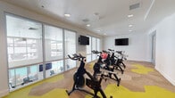 This single-floor spin studio is purpose-built for indoor cycling workouts, offering a clean, modern environment designed to motivate and energize. The room is furnished with a row of stationary cycling bikes arranged for an instructor-led class format, complemented by exercise accessories such as a stability ball and free weights. Two wall-mounted flat-screen televisions are strategically positioned at either end of the room, ensuring clear visibility from every bike. The space is defined by its bold, eye-catching carpet, which features a geometric hexagonal pattern in vibrant yellow and neutral tan tones, adding a dynamic visual energy to the floor. Recessed lighting in the textured ceiling provides consistent, even illumination throughout. Large floor-to-ceiling windows line one wall, flooding the studio with natural light and offering views of an outdoor pool and patio area, creating an open and airy atmosphere. The white walls remain clean and uncluttered, keeping the focus on the workout equipment. A glass partition wall along one side provides visual openness while maintaining a defined studio boundary. The overall ambiance is energetic yet functional, making this space well-suited for group fitness classes or individual cycling sessions.