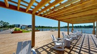Located along the Cape Fear River at 4126 River Rd in Wilmington, NC, this thoughtfully designed waterfront outdoor space offers an exceptional riverside experience. The elongated layout stretches along the water's edge, with every element oriented to maximize sweeping views of the river and its natural surroundings.

A wide wooden boardwalk serves as the primary pathway, connecting a series of distinct activity zones. The centerpiece is a generously sized artificial turf lawn, ideal for lawn games such as cornhole, with cable-railed fencing along the riverfront perimeter providing unobstructed sightlines to the water. Raised cedar planter beds run the length of the space, filled with ornamental grasses that add texture and a naturalistic aesthetic.

A covered pier extends directly over the river, fitted with Adirondack-style seating beneath a slatted pergola roof — a prime spot for relaxing and taking in the water views. An additional floating dock with mooring posts provides direct water access for boating. A utility structure, finished in a clean white shipping container design, houses electrical infrastructure and complements a nearby picnic area. An outdoor grill station adds to the functionality of the space.

The overall ambiance is open, breezy, and oriented around an active waterfront lifestyle, making this a well-equipped riverfront amenity destination.