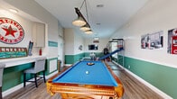 This dedicated game room is a well-appointed recreational space designed to deliver a full entertainment experience. The room's interior is defined by a two-tone wall treatment — a crisp cream upper half paired with a bold green wainscoting below — complemented by light wood-style flooring that runs the length of the space. Pendant and hanging lighting fixtures, including a stylish billiard-style light suspended above the centerpiece pool table, add character and ample illumination throughout.

The pool table, a handsome wood-framed model with a vibrant blue felt surface, anchors the room and invites competitive play. Along the walls, a variety of arcade-style games add to the entertainment lineup, including a skee-ball machine, a basketball shooting game complete with a ball return ramp, and a classic shuffleboard table. A wall-mounted flat-screen TV provides the perfect backdrop for watching the game while competing. Retro-themed wall art and vintage signage, including automotive-inspired décor, add personality and a nostalgic touch to the atmosphere.

Large windows along one side of the room allow natural light to fill the space, enhancing its open and lively feel. A glass-paned exterior door provides additional brightness and easy access. The overall ambiance is energetic and welcoming, making this game room a premier destination for leisure and friendly competition.