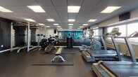 This fitness center at 112 Somerset St in New Brunswick, NJ, is a thoughtfully designed single-floor facility offering a broad range of workout options. The space features a drop-panel ceiling with recessed fluorescent lighting that ensures consistent, bright illumination throughout. Dark rubber flooring covers the entire area, providing durability and safety during exercise.

The cardio zone is lined with multiple treadmills and elliptical machines positioned along a windowed wall, allowing natural light to filter in while offering views of the surrounding area. Large wall mirrors enhance the sense of space and allow users to monitor their form. Mounted flat-screen televisions are positioned for easy viewing during workouts.

The strength training section includes a cable machine, a power rack with barbells and weight plates, a flat adjustable bench, and a full dumbbell rack. A vibrant blue accent wall with framed artwork adds personality to the functional training area, which is stocked with medicine balls on a vertical rack, stability balls, foam rollers, and resistance tools.

A utility area features a water cooler station with a wall-mounted television and accessory storage shelf, rounding out the amenities. The overall ambiance is clean, modern, and functional, catering to a variety of fitness needs.