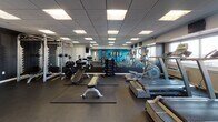 This fitness center at 112 Somerset St in New Brunswick, NJ, is a thoughtfully designed single-floor facility offering a broad range of workout options. The space features a drop-panel ceiling with recessed fluorescent lighting that ensures consistent, bright illumination throughout. Dark rubber flooring covers the entire area, providing durability and safety during exercise.

The cardio zone is lined with multiple treadmills and elliptical machines positioned along a windowed wall, allowing natural light to filter in while offering views of the surrounding area. Large wall mirrors enhance the sense of space and allow users to monitor their form. Mounted flat-screen televisions are positioned for easy viewing during workouts.

The strength training section includes a cable machine, a power rack with barbells and weight plates, a flat adjustable bench, and a full dumbbell rack. A vibrant blue accent wall with framed artwork adds personality to the functional training area, which is stocked with medicine balls on a vertical rack, stability balls, foam rollers, and resistance tools.

A utility area features a water cooler station with a wall-mounted television and accessory storage shelf, rounding out the amenities. The overall ambiance is clean, modern, and functional, catering to a variety of fitness needs.