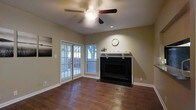 This single-floor residence offers a comfortable and well-appointed living space designed with both style and function in mind. The living room serves as the heart of the home, showcasing rich, dark hardwood flooring that adds warmth and character to the space. A classic fireplace with a black surround and a clean white mantel serves as a striking focal point, providing both aesthetic appeal and a cozy atmosphere. Above, a ceiling fan with an integrated light fixture ensures year-round comfort and ambient lighting. Natural light flows generously into the room through a set of double windows and a glass-paneled door, which appears to open onto an outdoor deck or patio area, offering a seamless connection to the exterior. The neutral, warm-toned walls complement the overall palette of the space, creating a cohesive and inviting ambiance. White baseboards and trim provide clean, crisp contrast throughout. The layout is open and unobstructed, allowing for flexible furniture arrangements to suit a variety of living styles. The textured ceiling adds a subtle traditional detail, rounding out this well-maintained, move-in ready home.